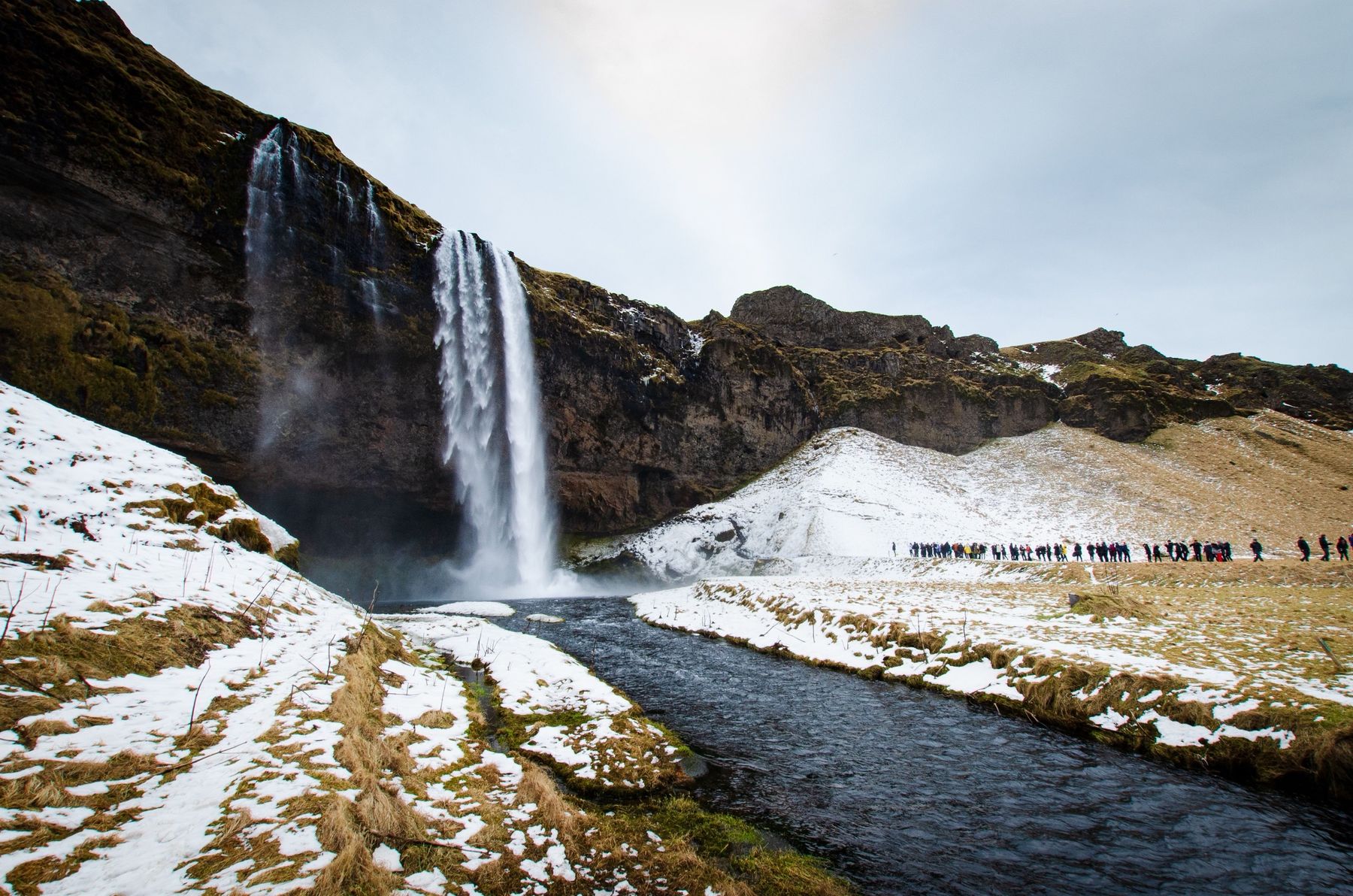 Seljalandsfoss waterfall in winter, with snow on the slopes and a line of walkers on the path