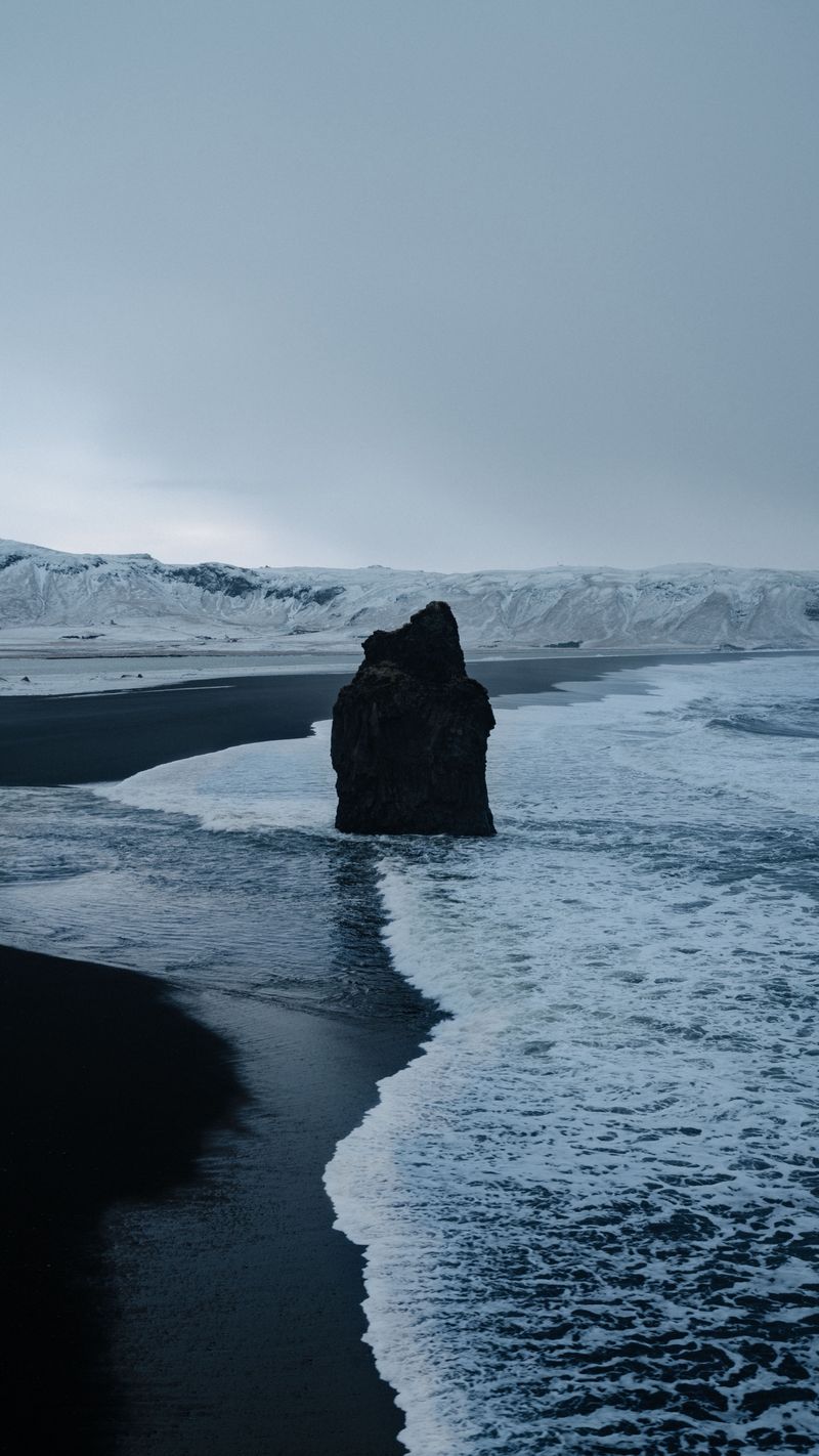 A sea stack in the surf off a black sand beach with snowy cliffs behind