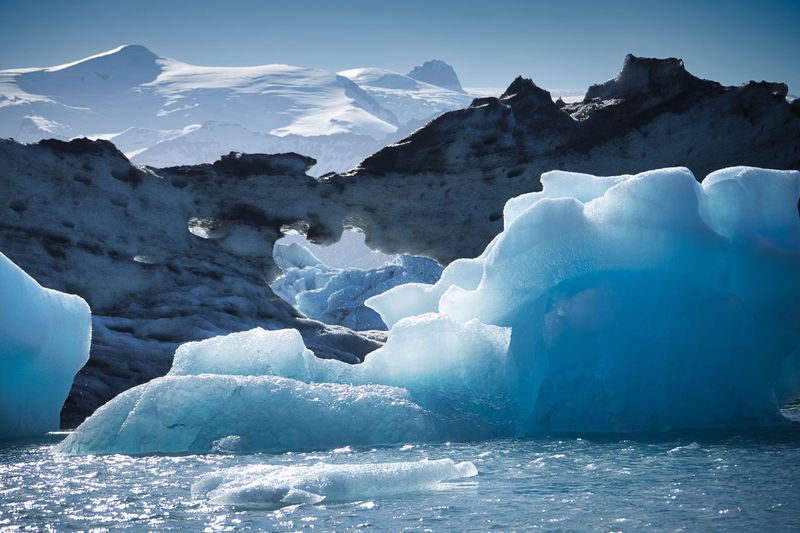 A glowing blue iceberg in front of dark ash-streaked ice and snowy peaks