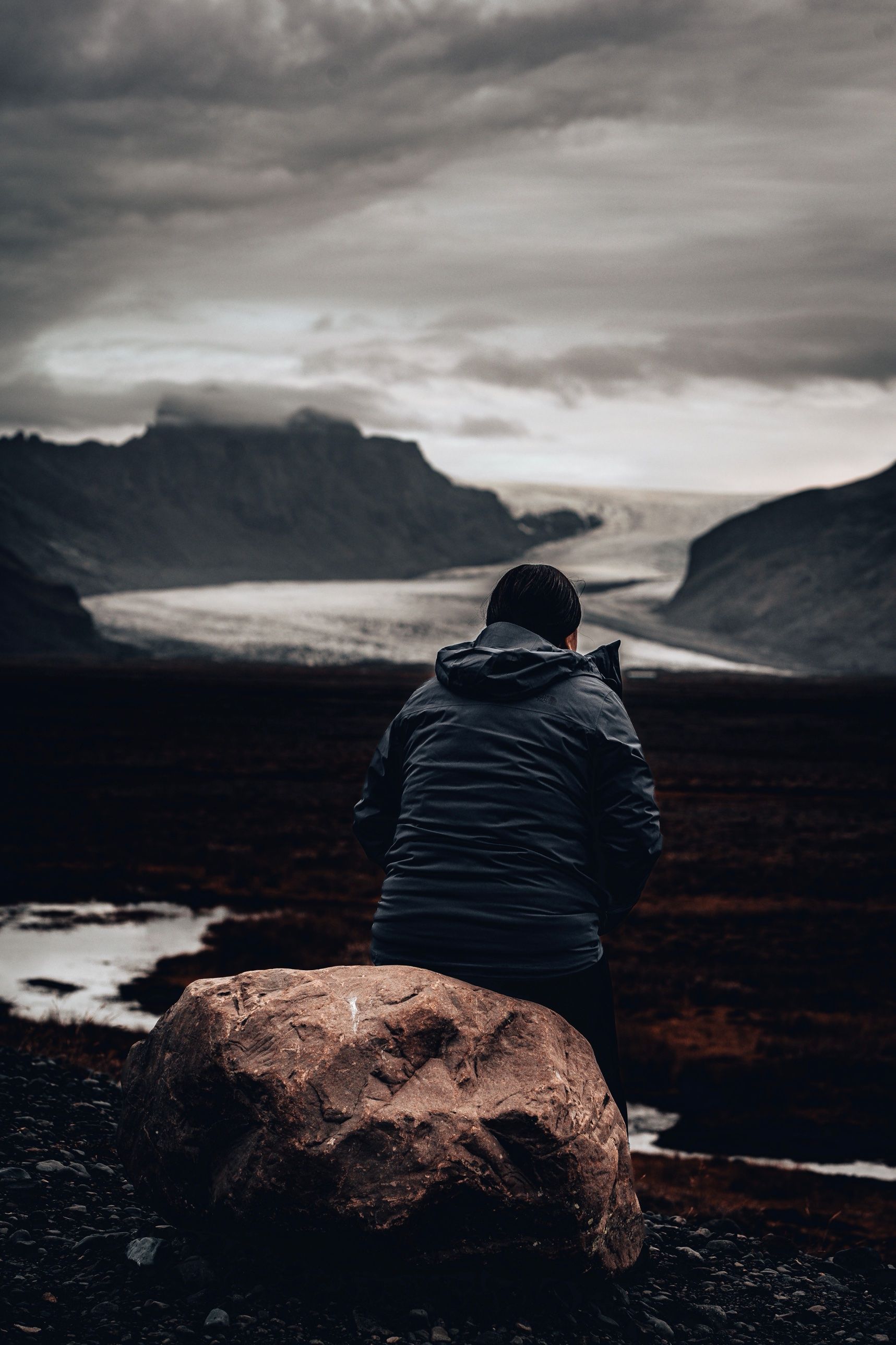 A walker sitting by a boulder looking out at a glacier tongue between dark mountains