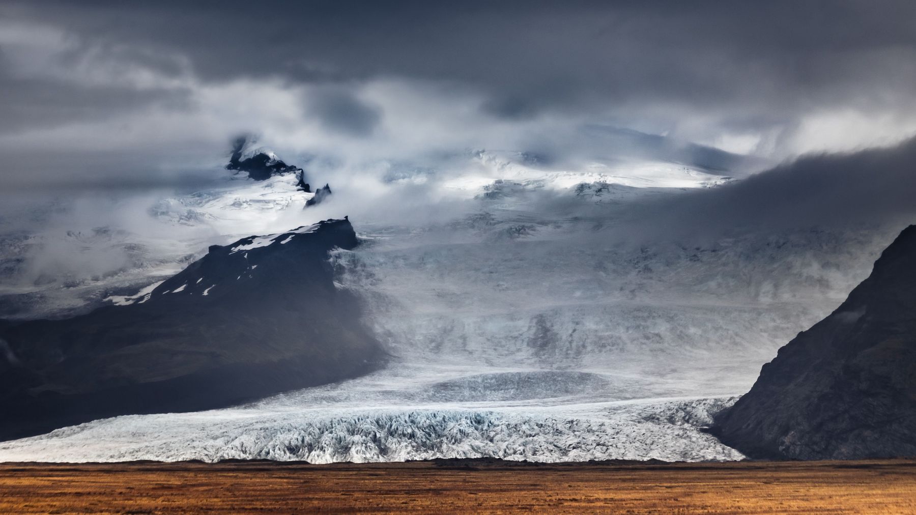A glacier spilling from cloud-covered mountains above golden grassland