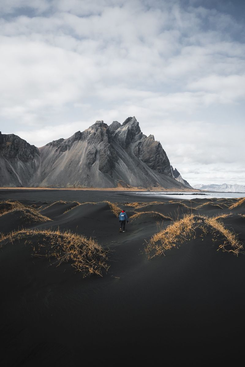 A walker in a blue jacket crossing black dunes below the jagged peaks of Vestrahorn