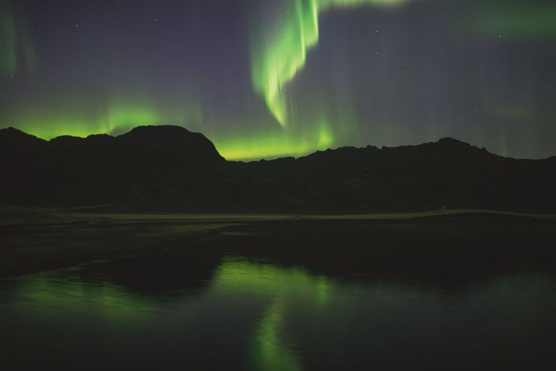 Green northern lights over dark mountains, reflected in a still lake