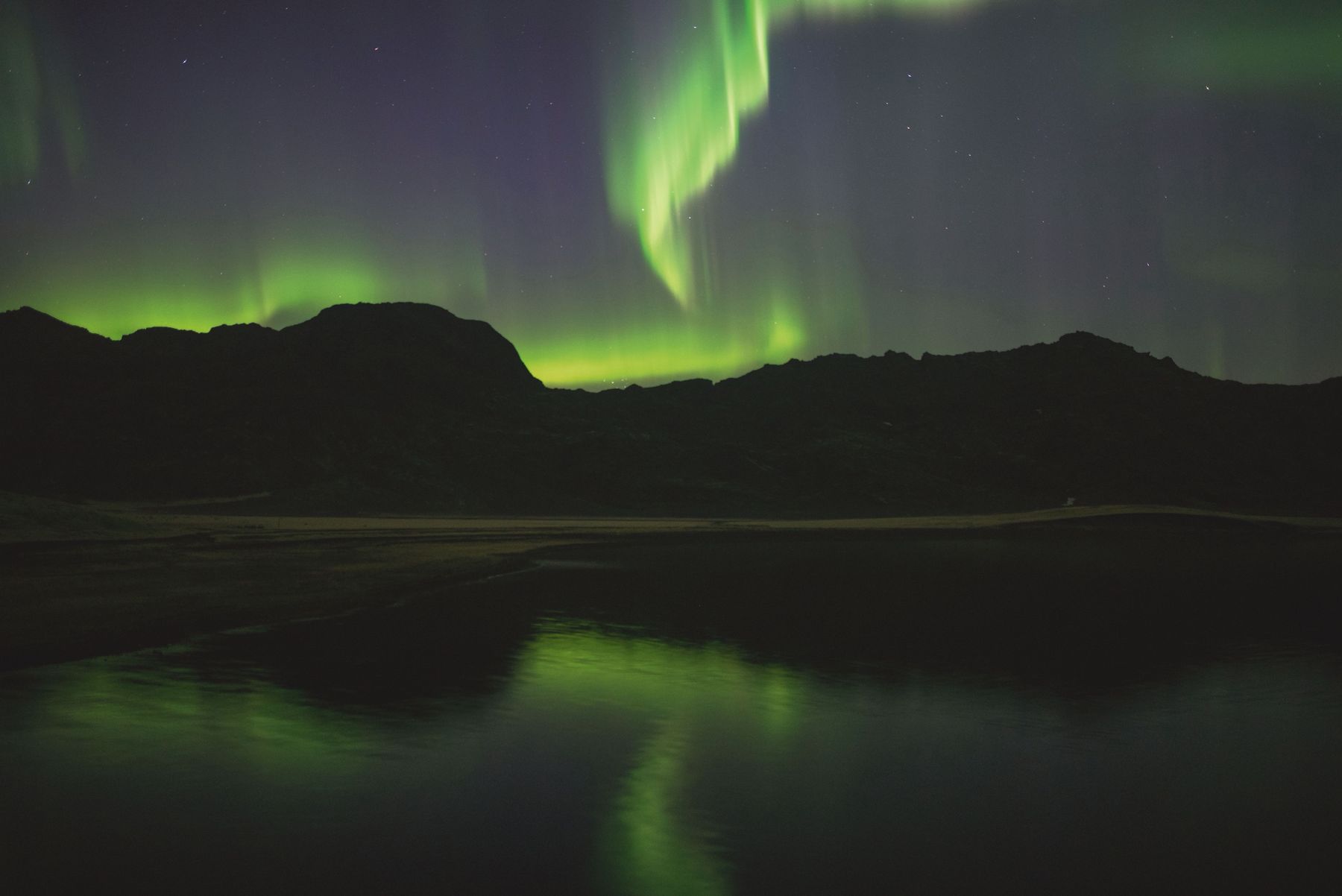Green northern lights over dark mountains reflected in a lake