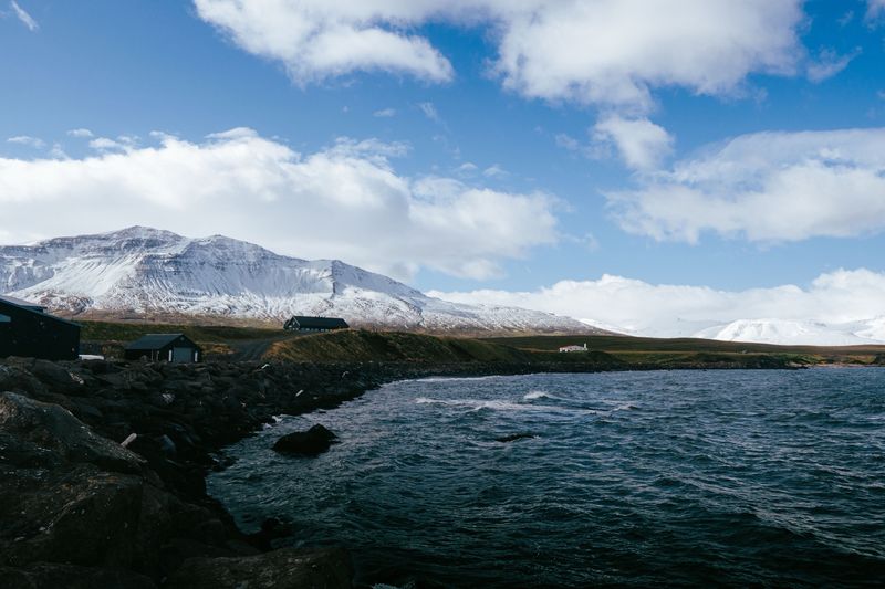 Small dark houses on a rocky shore below a snow-capped mountain under blue sky